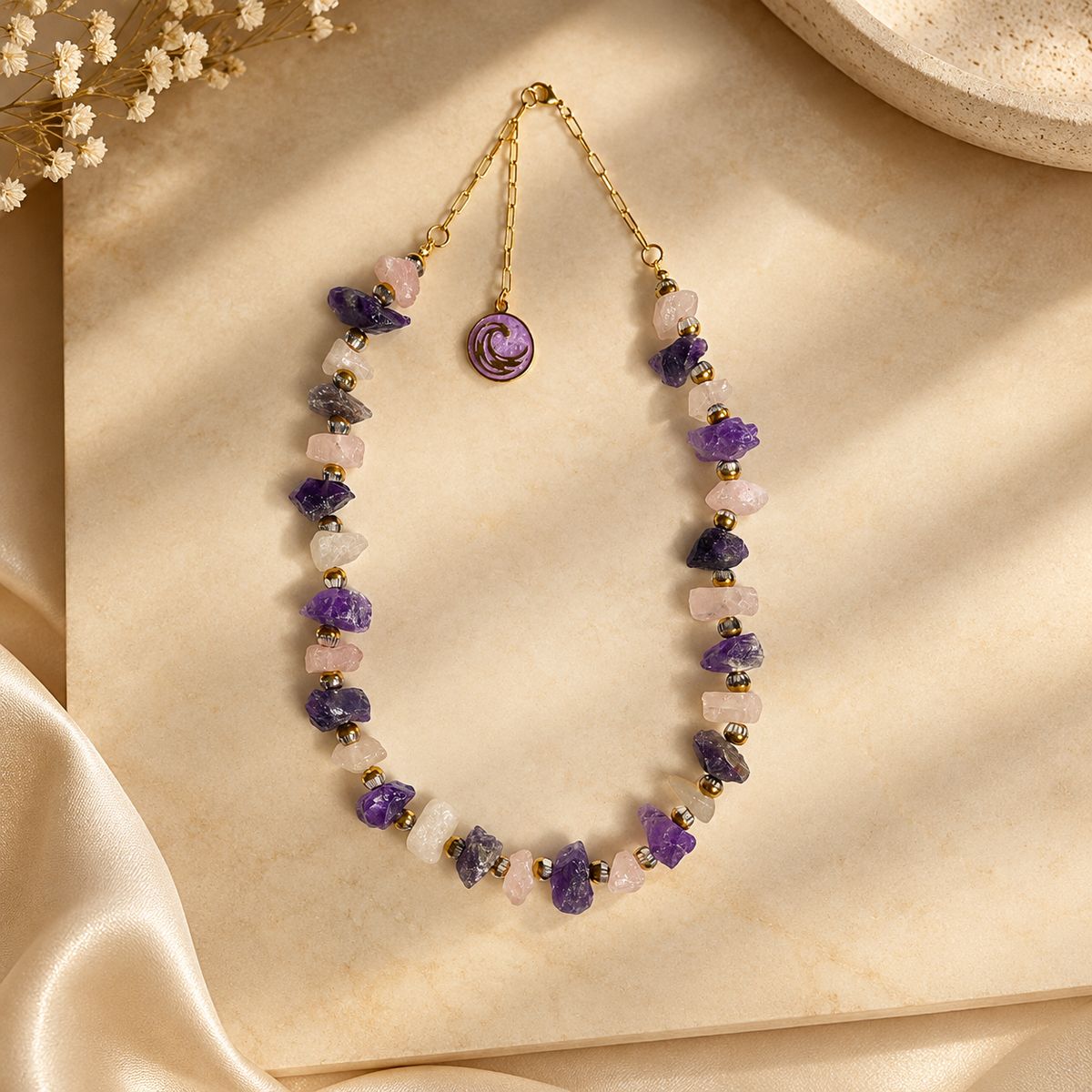 A handmade necklace made of raw purple, pink, and white gemstone chips with a gold chain and a rose-shaped charm, displayed on a beige textured surface with decorative dried flowers and satin fabric.