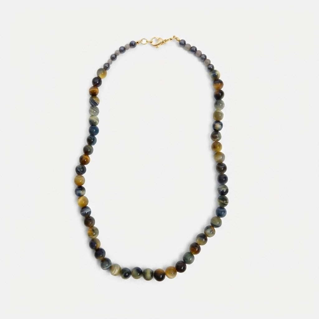 A beaded necklace made of polished round gemstone beads in earthy brown, gray, and blue tones, with a small gold clasp, shown on a light gray background.