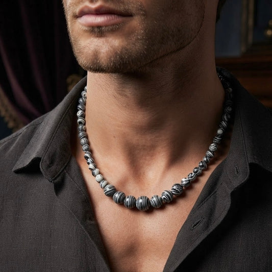 A close-up of a man wearing a chunky beaded necklace made of alternating black and silver metallic beads, styled with a dark button-down shirt.