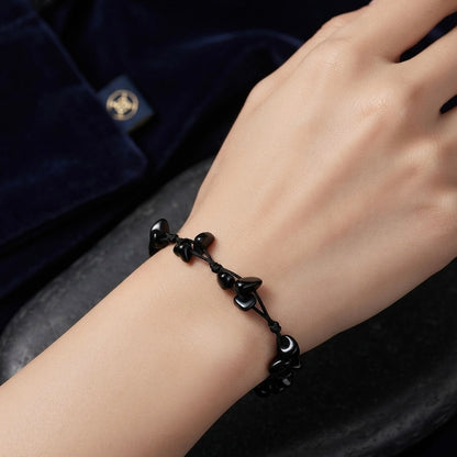 A close-up of a person's wrist wearing a black beaded cord bracelet with small polished black beads and a central black heart-shaped bead. The background includes a dark velvet garment with a gold emblem.