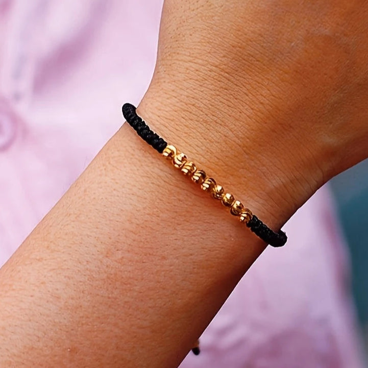 Close-up of a wrist wearing a black and gold beaded bracelet with a blurred pink shirt background.