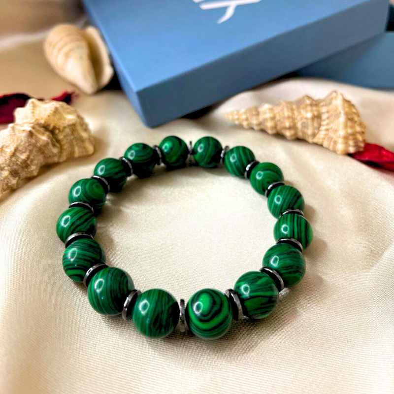 Green beaded bracelet on a sandy surface with seashells and a blue box in the background.