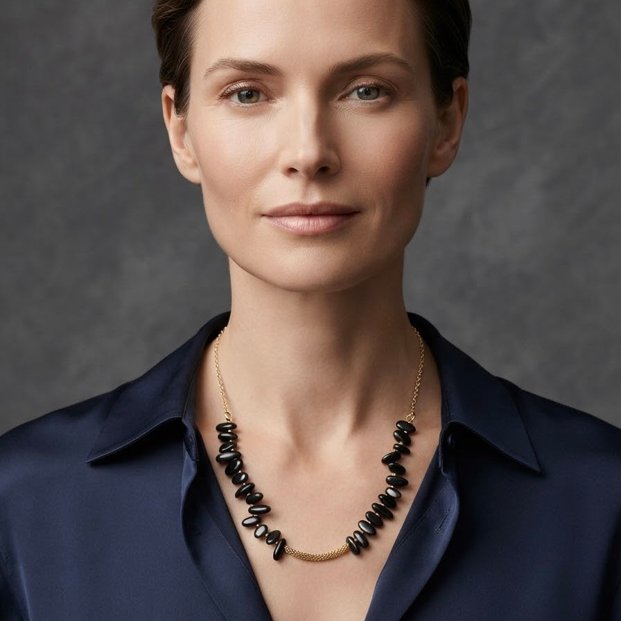 A close-up portrait of a woman wearing a gold necklace with irregular black beads and a small gold heart pendant, paired with a dark navy satin shirt against a textured gray background.