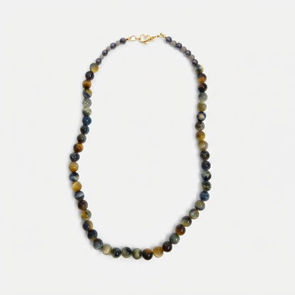 A beaded necklace made of polished round gemstone beads in earthy brown, gray, and blue tones, with a small gold clasp, shown on a light gray background.