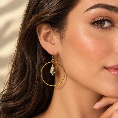 A close-up of a woman wearing a gold hoop earring with a small seashell charm, highlighting the jewelry against her dark brown hair and warm skin tones.