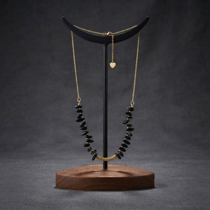 A gold chain necklace with black stone beads draped over a black crescent moon stand on a round wooden base, set against a dark gray background.